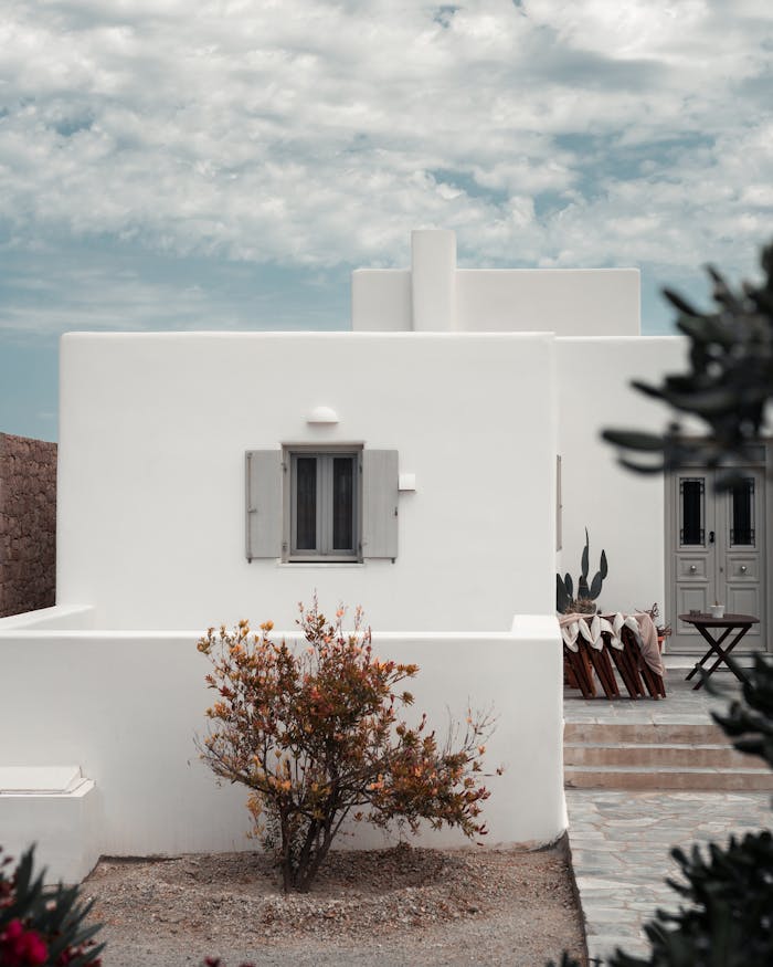 about-01 Whitewashed house with wooden shutters and garden in a tranquil rural setting under a cloudy sky.